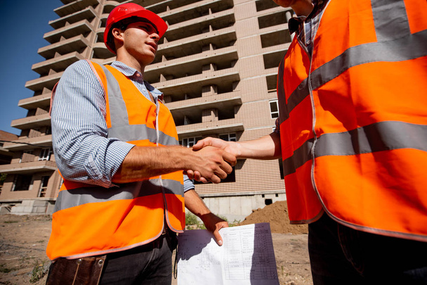Two men dressed in shirts, orange work vests and helmets shake hands against the background of a multistorey building - 写真・画像