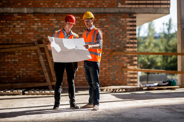 Architect  and construction manager dressed in orange work vests and helmets discuss documentation on a brick wall background - Foto, Bild