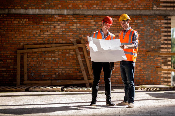 Structural engineer and architect dressed in orange work vests and helmets discuss documentation on a brick wall background - Photo, Image