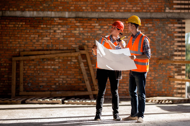 Structural engineer and architect dressed in orange work vests and helmets discuss documentation on a brick wall background - Foto, Bild