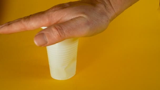 A mans hand crushes a white disposable plastic cup on a yellow background, footage ideal for topics such as ecology and recycling - Imágenes, Vídeo