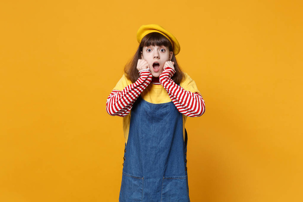 Šokovaná Dívka teenager v francouzský baret, letní Džínové šaty, držet ústa dokořán, strčil ruce na tváře izolované na pozadí žluté zdi. Lidé upřímné emoce životní styl koncept. Vysmívat se kopie prostor - Fotografie, Obrázek