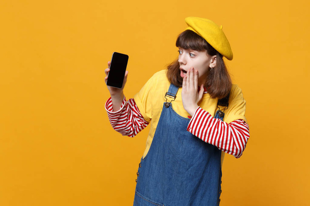 Shocked girl teenager in french beret put hand on face, holding mobile phone with blank empty screen isolated on yellow wall background. People sincere emotions, lifestyle concept. Mock up copy space - Photo, image