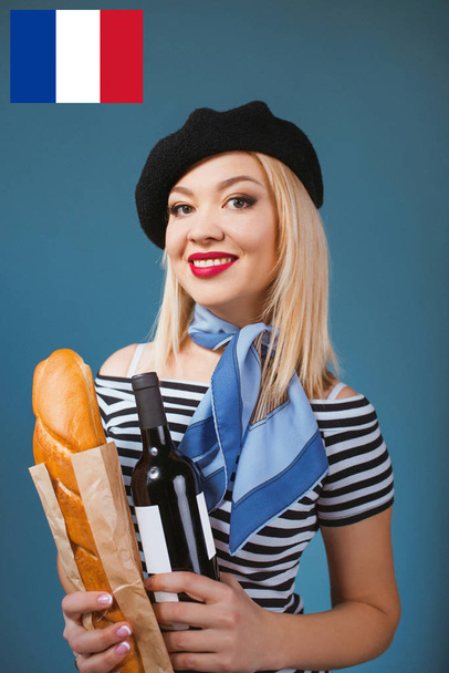 portrait of beautiful blonde french woman in beret, scarf, back and white shirt, with bottle of wine and bread baguette in her arms with French flag on background - Photo, Image