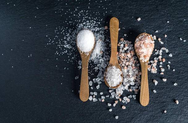 Different types of salt. Sea, himalayan and kitchen salt. Top view on three wooden spoons on black background - Zdjęcie, obraz