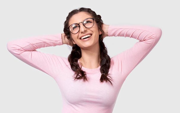 Positive Caucasian woman student keeps hands on head, smiling broadly, wears pink casual clothes and round spectacles, isolated over white background. Happy young woman with braids hairstyle on white - Photo, Image