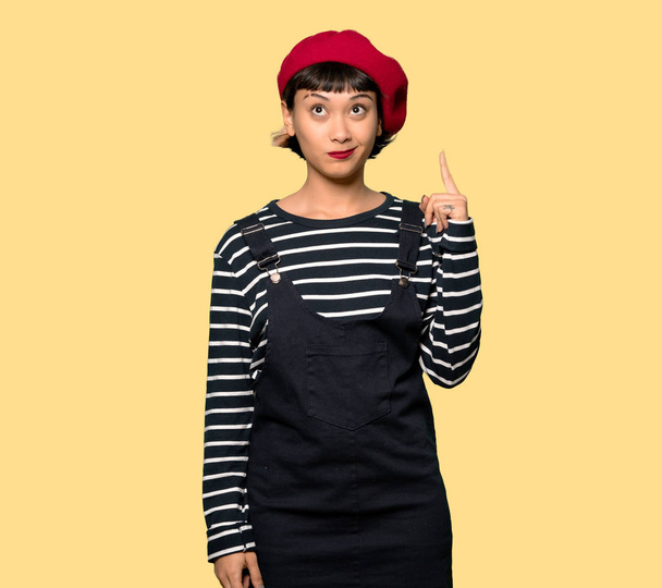 Young woman with beret pointing with the index finger a great idea over yellow background - 写真・画像