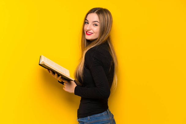 Joven bonita mujer sobre fondo amarillo sosteniendo un libro y disfrutando de la lectura
 - Foto, Imagen