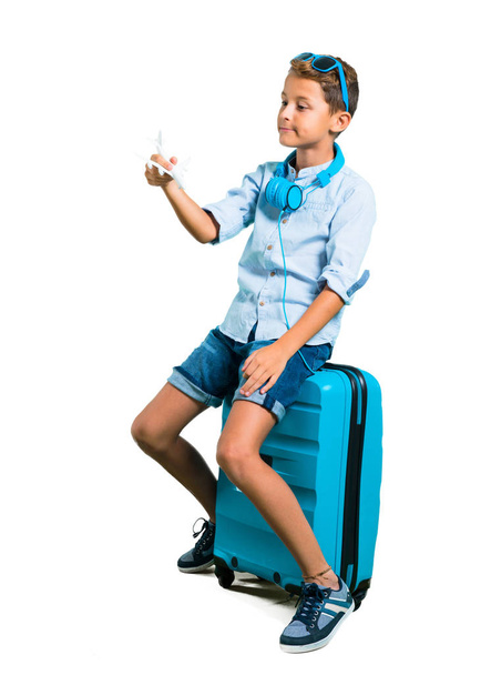 Повне тіло хлопчика з сонцезахисними окулярами та навушниками, що подорожують зі своєю валізою, тримаючи іграшковий літак на ізольованому білому тлі
 - Фото, зображення