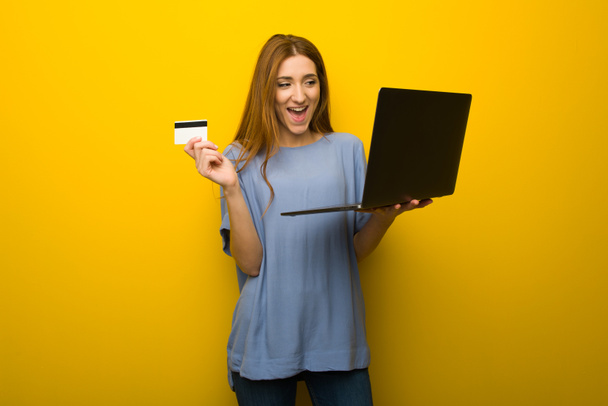 Chica pelirroja joven sobre fondo amarillo de la pared con ordenador portátil y tarjeta de crédito
 - Foto, imagen