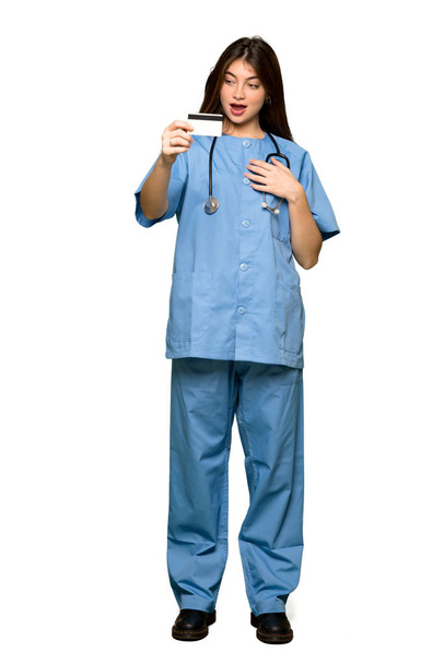 Full-length shot of Young nurse holding a credit card and surprised on isolated white background - Valokuva, kuva