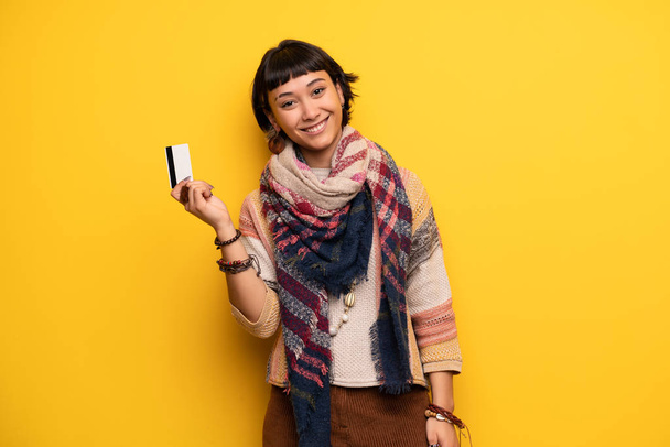 Joven hippie mujer sobre amarillo pared celebración de una tarjeta de crédito
 - Foto, Imagen