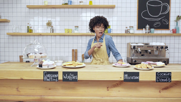 piękny uśmiechający się właściciel african american kobieta café w fartuch taniec za ladą z deserów - Materiał filmowy, wideo