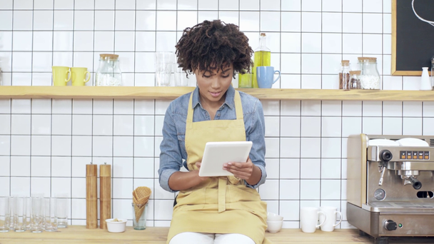 mooie lachende Afro-Amerikaanse vrouwelijke kassier zittend op de teller en met behulp van digitale tablet in café  - Video