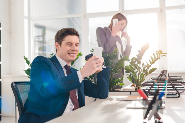 selective focus of cheerful businessman holding smartphone and watching video near female coworker on background - Foto, Bild