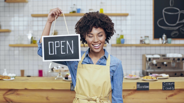 beautiful smiling african american female cafe owner in apron holding open sign and looking at camera - Video