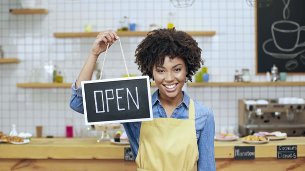 beautiful african american female cafe owner in apron holding open sign and looking at camera - Footage, Video