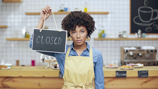 beautiful african american female cafe owner in apron holding closed sign and looking at camera - Footage, Video