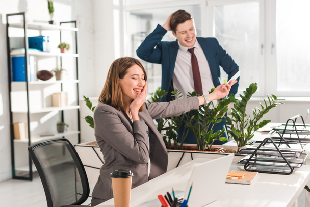 happy businesswoman touching face whle holding smartphone and laughing with coworker in office  - Foto, Bild