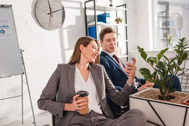 привлекательная деловая женщина улыбается, держа в руках бумажную чашку и смартфон рядом с коллегой в офисе
  - Фото, изображение