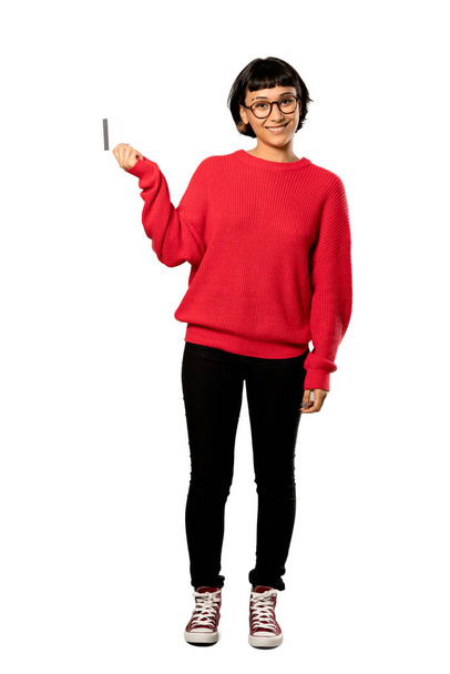 クレジット カードを持っていると考えて分離の白い背景の上に赤いセーターと短い髪の女の子のフルレングス ショット - 写真・画像