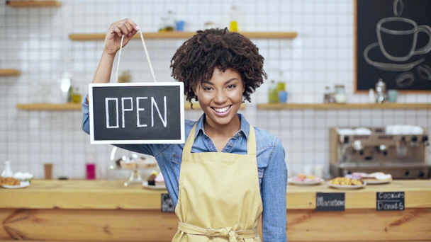 pomalý pohyb africké americké krásné ženské kavárník v zástěře hospodářství otevřené znamení a při pohledu na fotoaparát - Záběry, video