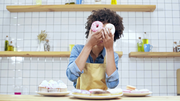 de eigenaar van de prachtige Afrikaanse Amerikaanse vrouwelijke café in schort holding donuts voor gezicht, lachen en kijken naar camera - Video
