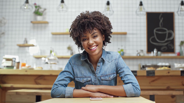 όμορφη αφρικανική αμερικανική γυναίκα πελάτη σε τζιν μπλούζα βλέπουν φωτογραφική μηχανή και να χαμογελά στο café - Πλάνα, βίντεο