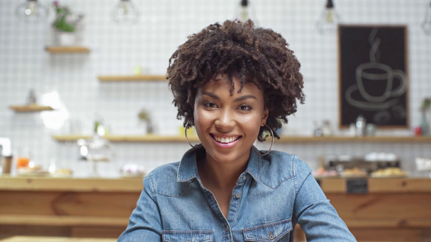 beautiful african american female customer in denim blouse looking at camera and smiling in cafe - Filmati, video
