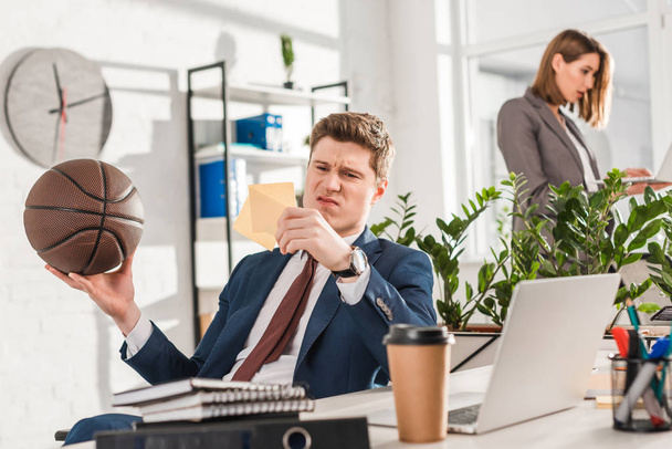 selective focus of dissatisfied businessman looking at sticky notes while holding basketball in office with coworker on background, procrastination concept - Фото, изображение