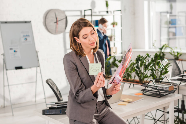 selective focus of businesswoman holding sticky note with later lettering and magazine in hands with coworker on background, procrastination concept - Fotografie, Obrázek