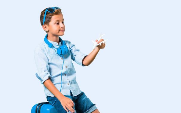 Хлопчик з сонцезахисними окулярами та навушниками, що подорожують зі своєю валізою, тримаючи іграшковий літак на синьому фоні
 - Фото, зображення