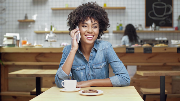 hermosa sonriente afroamericana cliente femenino sentado en la mesa con pastelería y café mientras habla en el teléfono inteligente en la cafetería
 - Metraje, vídeo