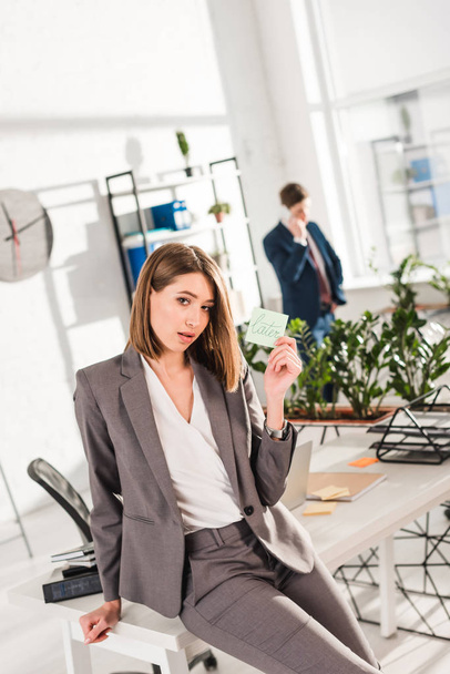 selective focus of businesswoman holding sticky note with later lettering with coworker on background, procrastination concept - Фото, изображение