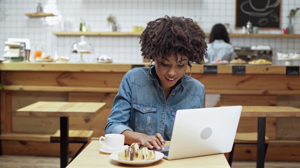 piękne african american kobieta freelancer siedząc przy stole, picie kawy, uśmiechnięci i przy użyciu laptopa w kawiarni - Materiał filmowy, wideo