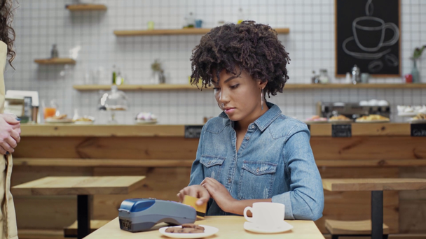 cajero femenino que sostiene terminal mientras que el cliente femenino afroamericano hermoso que paga con tarjeta de crédito en la cafetería
 - Metraje, vídeo