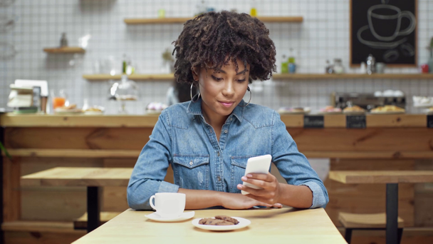 schöne afrikanisch-amerikanische Kundin sitzt, trinkt Kaffee, benutzt Smartphone und lächelt im Café - Filmmaterial, Video
