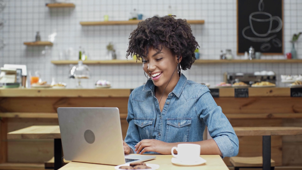 piękny szczęśliwy african american kobieta freelancer w słuchawki za pomocą laptopa i słuchanie muzyki w kawiarni - Materiał filmowy, wideo