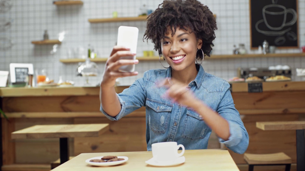 güzel Afrikalı-Amerikalı kadın müşteri alarak selfie gösteren barış işareti süre gülümseyen ve hava üfleme kafede öp  - Video, Çekim