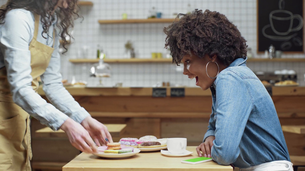 číšnice přináší koblihy, košíčky a macarons na krásném úsměvu afroamerické ženy zákazníka v kavárně - Záběry, video