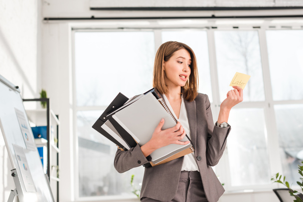 привлекательная деловая женщина, держащая папки и глядя на липкую ноту с более поздними надписями в офисе
  - Фото, изображение