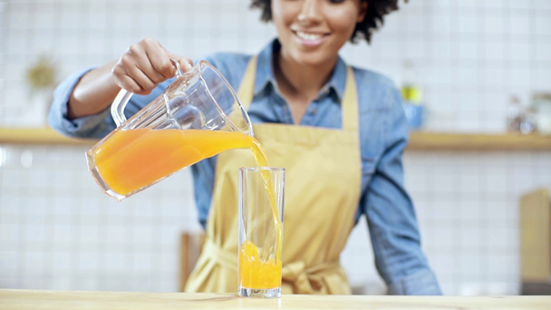 smiling african american female cafe owner in apron putting glass on counter and pouring orange juice - Footage, Video