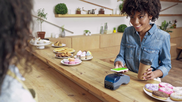 hermosa sonriente afroamericana cliente femenino pagando con tecnología nfc usando teléfono inteligente en la cafetería
 - Imágenes, Vídeo