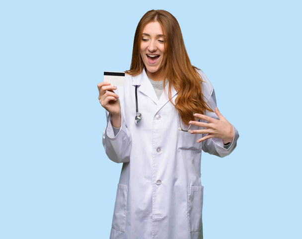 Redhead doctor woman holding a credit card and surprised on isolated blue background - Φωτογραφία, εικόνα
