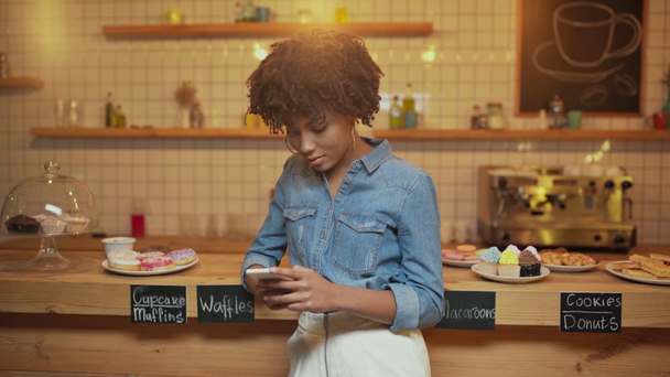 enfoque selectivo de la hermosa afroamericana propietaria de café femenino utilizando teléfono inteligente cerca del mostrador con pastelería en la cafetería
 - Imágenes, Vídeo