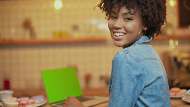 επιλεκτική εστίαση του όμορφη χαμογελαστή αφρικανική αμερικανική freelancer χρησιμοποιώντας φορητό υπολογιστή με κενή οθόνη στο café  - Πλάνα, βίντεο