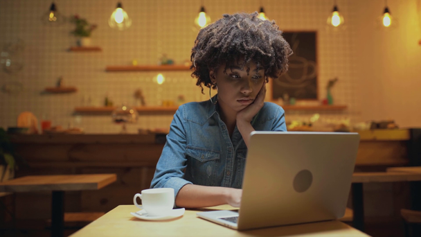 beautiful bored african american female freelancer sitting at table, drinking coffee and using laptop in cafe - Footage, Video