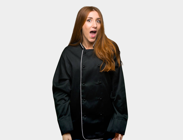 Joven chef pelirroja mujer con sorpresa y expresión facial impactada sobre fondo gris aislado
 - Foto, imagen