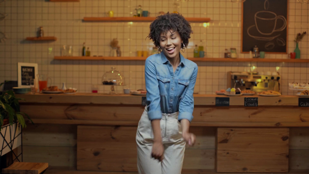красивая африканская владелица кафе в джинсовой блузке танцует, улыбается и смотрит в камеру в кафе
 - Кадры, видео