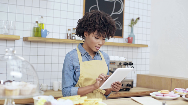 prachtige Afrikaanse Amerikaanse vrouwelijke kassa met behulp van digitale tablet achter balie met desserts in café  - Video